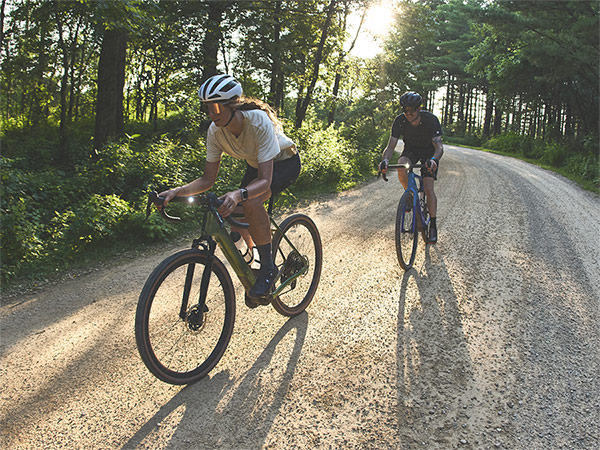 Cyclists wearing helmets while riding bikes on tree-lined gravel path with daytime running lights on