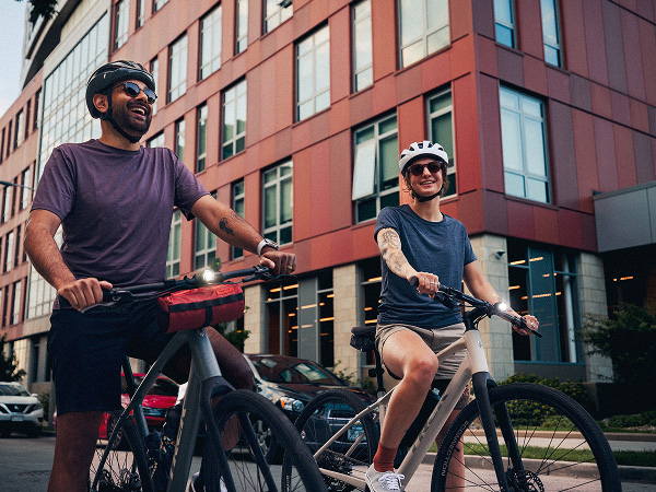 City buildings behind cyclists wearing helmets while standing with bikes and daytime running lights turned on