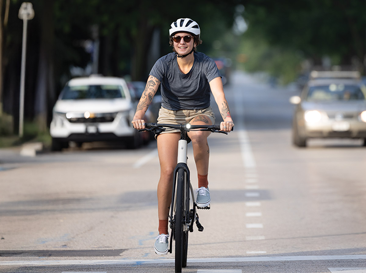 Cyclist wearing helmet and sunglasses while riding bike on paved city road with daytime running light turned on and cars in background