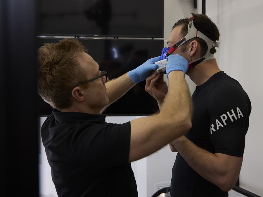 A person wearing gloves adjusts a respiratory mask on another person dressed in a Rapha shirt in an indoor setting