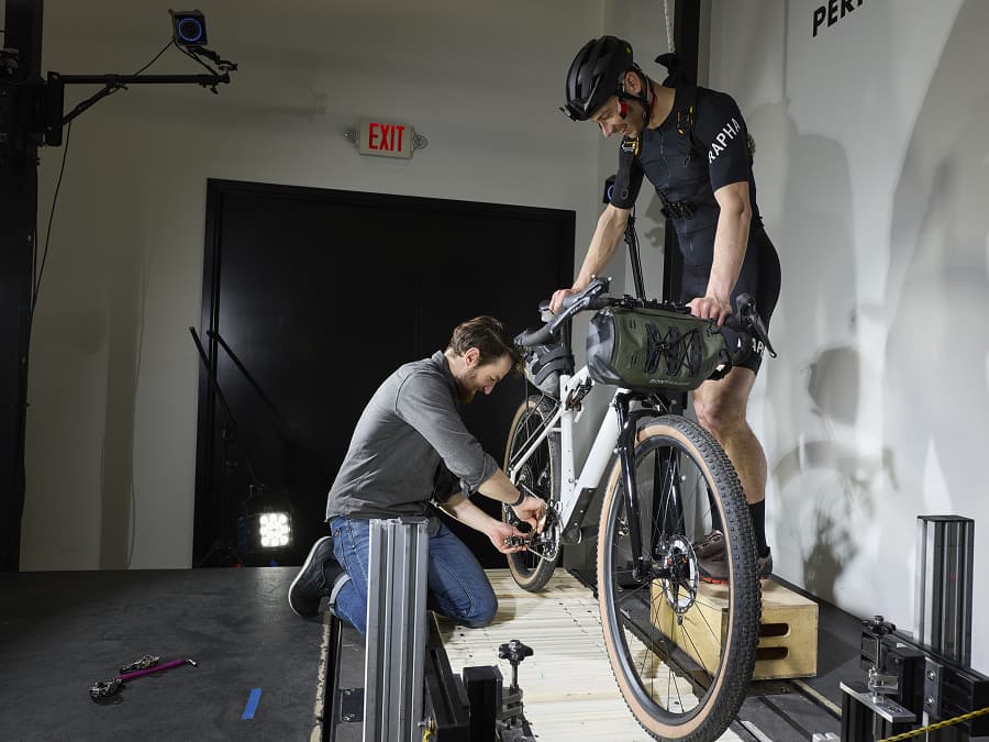 A person kneels to adjust the front wheel of a stationary bike, while another person wearing a helmet and cycling gear stands on the bike.