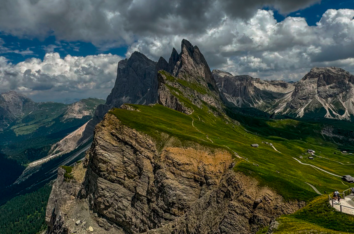 Sharp, jagged mountain peaks rise above green slopes and dramatic cliffs under a cloudy sky, with a few cyclists visible on a path in the lower right corner