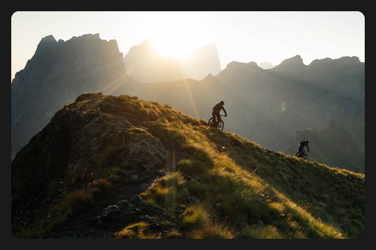 Cyclists wearing helmets while riding bikes on mountain trail with sun shining over mountains in background