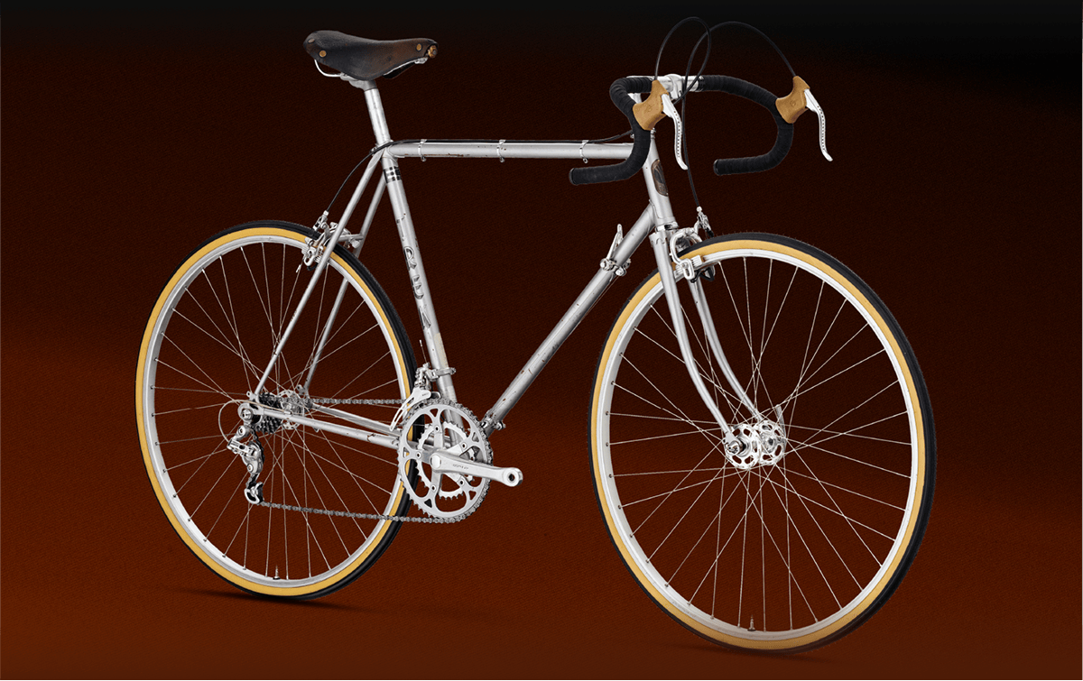 A silver vintage road bicycle with drop handlebars, brown saddle, and tan-walled tires is displayed against a dark brown background