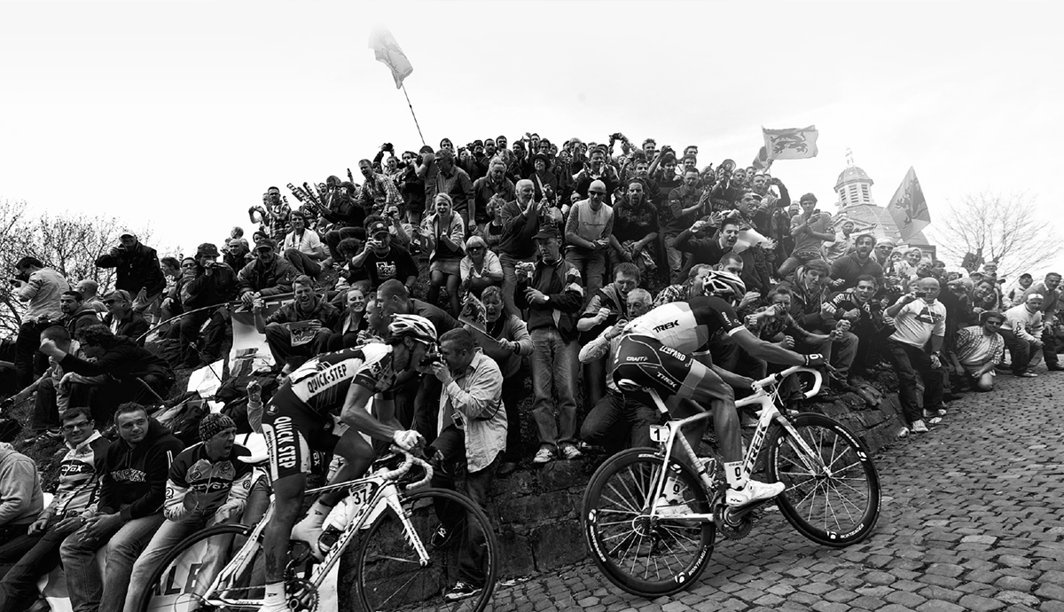Cyclists ride up a cobblestone street during a race as a large crowd sits and stands closely together on a stone embankment, cheering and watching
