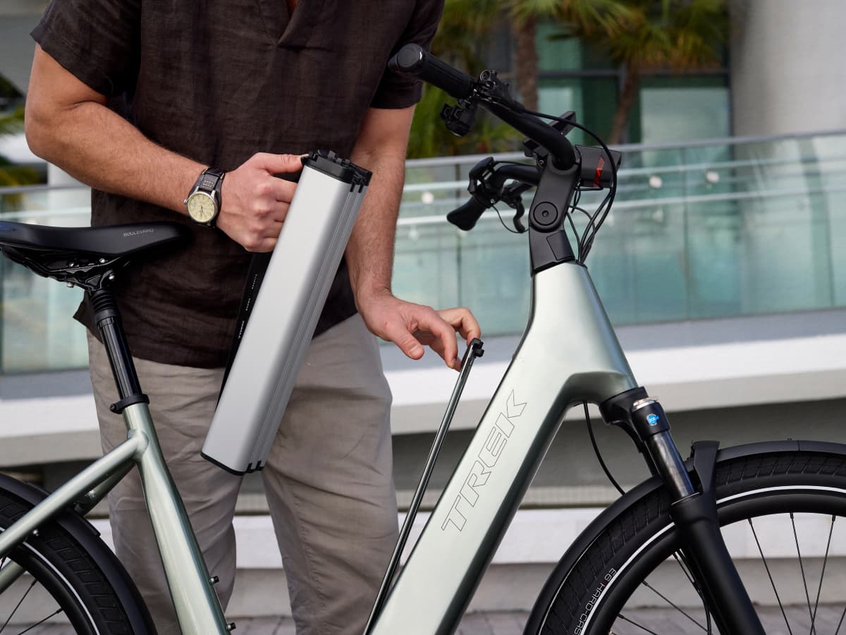 A person removes the battery from a silver Trek Charter+ bike parked outdoors, with a modern glass building in the background.