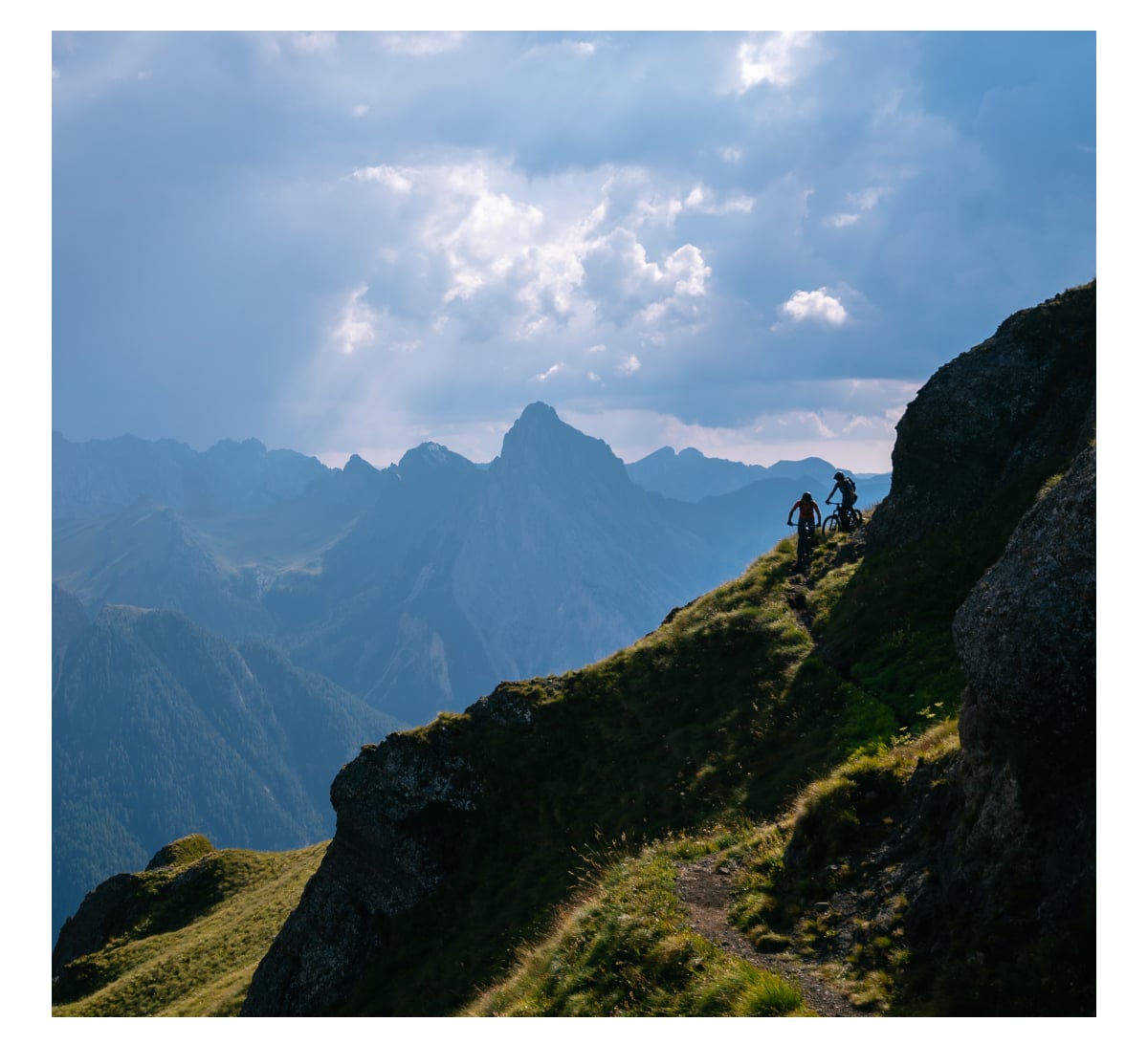 Two silhouetted mountain bikers ride along a narrow, grassy trail on a steep mountainside with dramatic, cloudy skies and layered mountain ranges in the background.