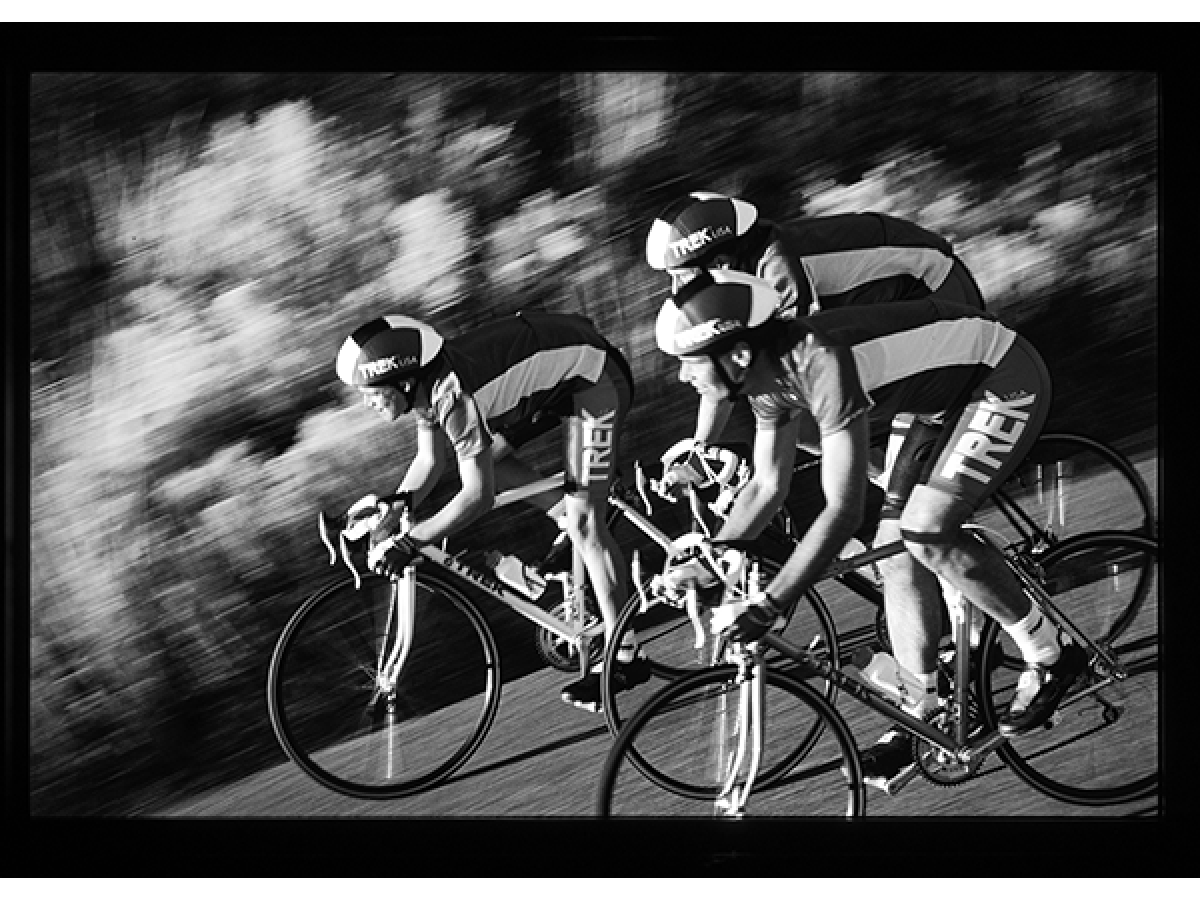 Alt text: Three cyclists in Trek kits and aero helmets ride tightly paced in a black-and-white motion-blurred action shot.