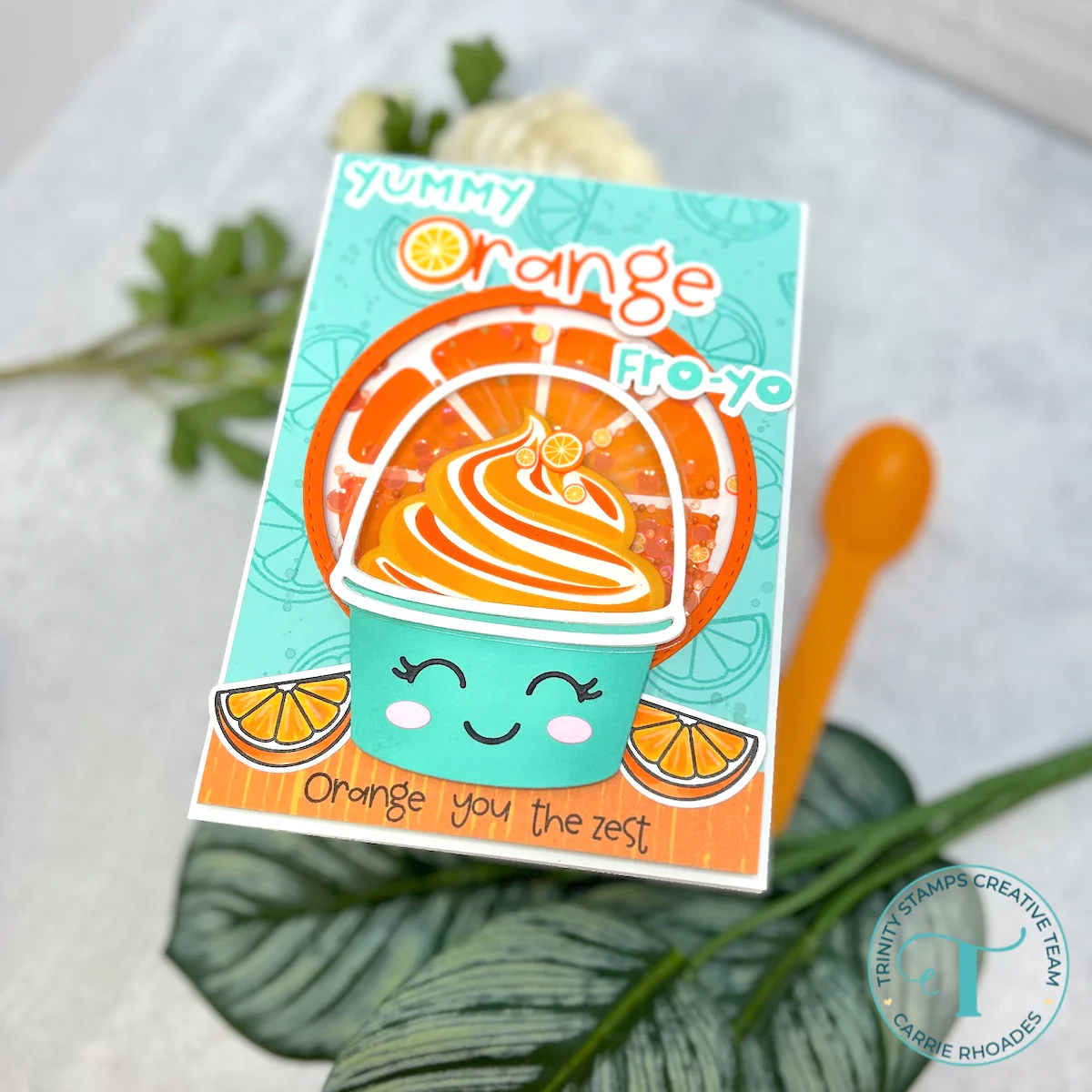 Orange swirl frozen yogurt in a teal cup with a smiley face. Decorated with orange wedges and an orange background with shaker elements. Reads "yummy orange fro-yo / Orange you the zest"
