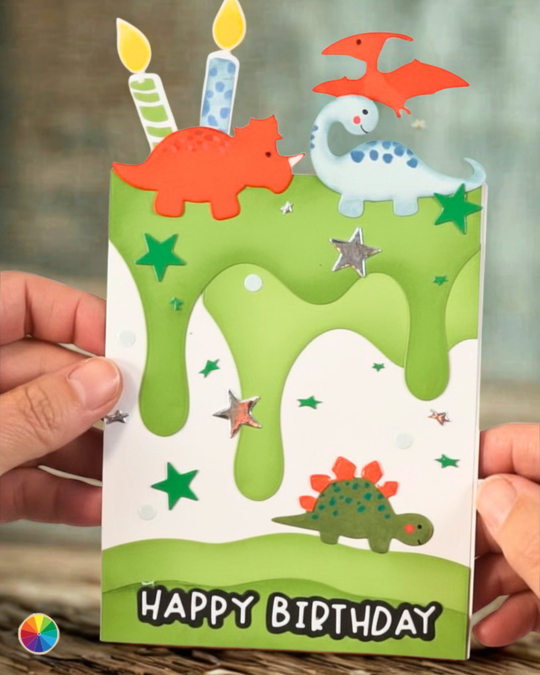 Prehistoric Pals dino cutouts top a layered paper cake dripping with green icing and covered in stars. Two candles indicate this is for a 2-year-old with a message reading "Happy Birthday."
