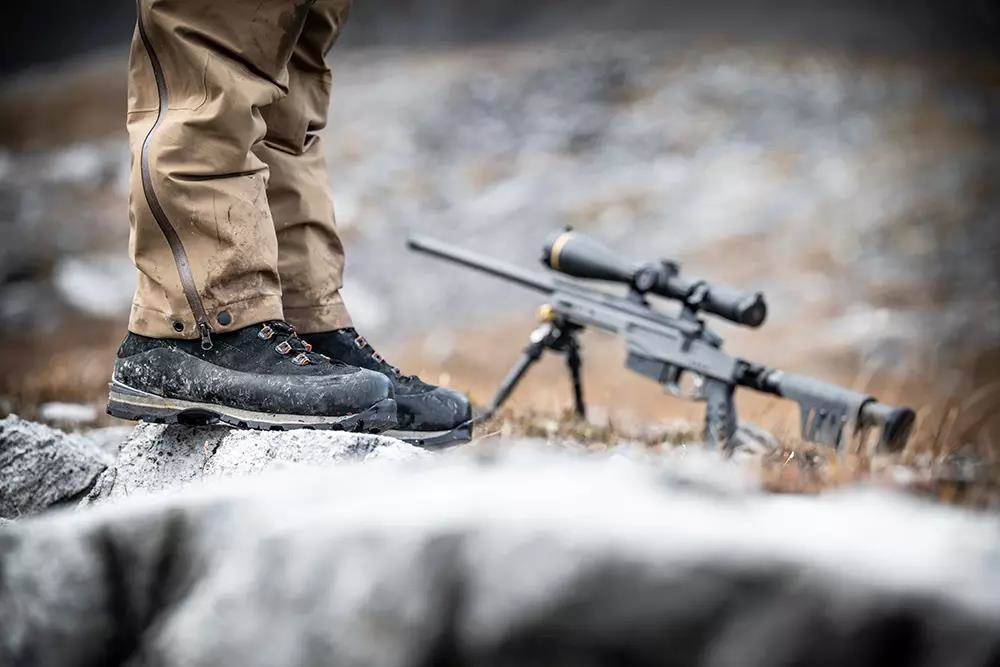 Legs of a hunter standing on a rock in front of a large black rifle with scope