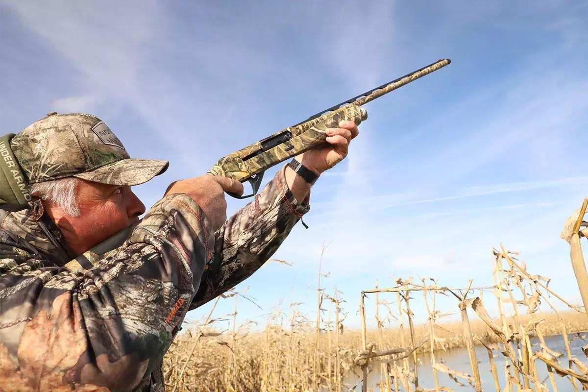 Hunter in brown camo taking aim in tall grass with rifle