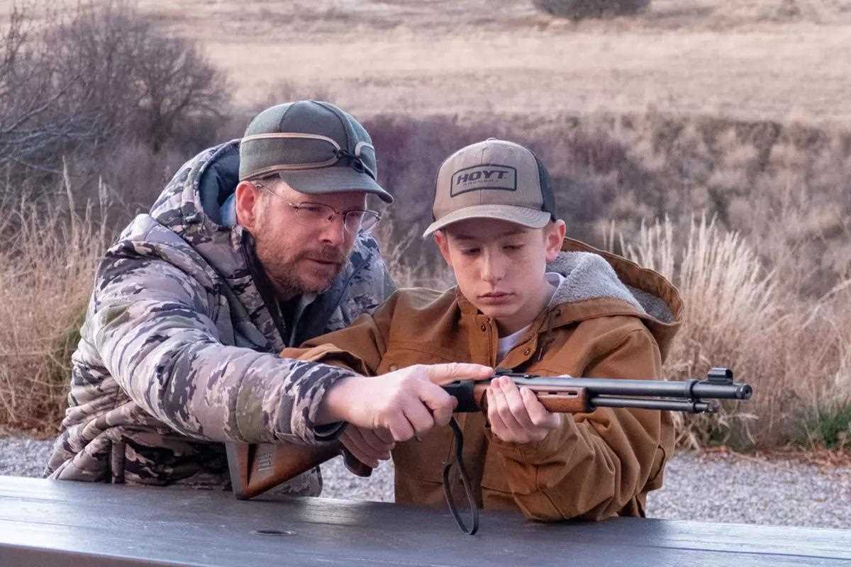 Man explaining to a young man the details of a rifle; brown field behind them