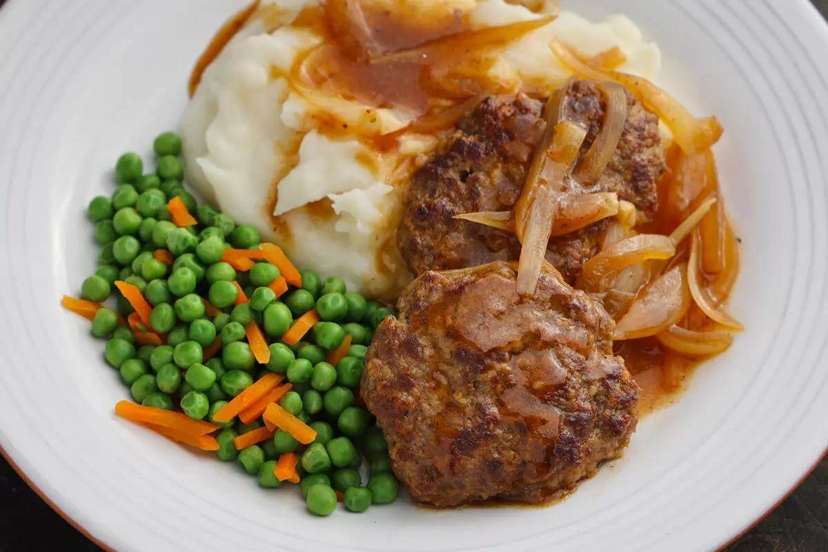 Round white plate with two Salisbury steaks with brown gravy & cooked onion on top, mashed potatoes and brown gravy, and peas & carrots beside it