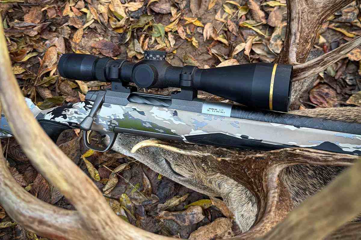 Camo rifle with black riflescope laying on wooded ground
