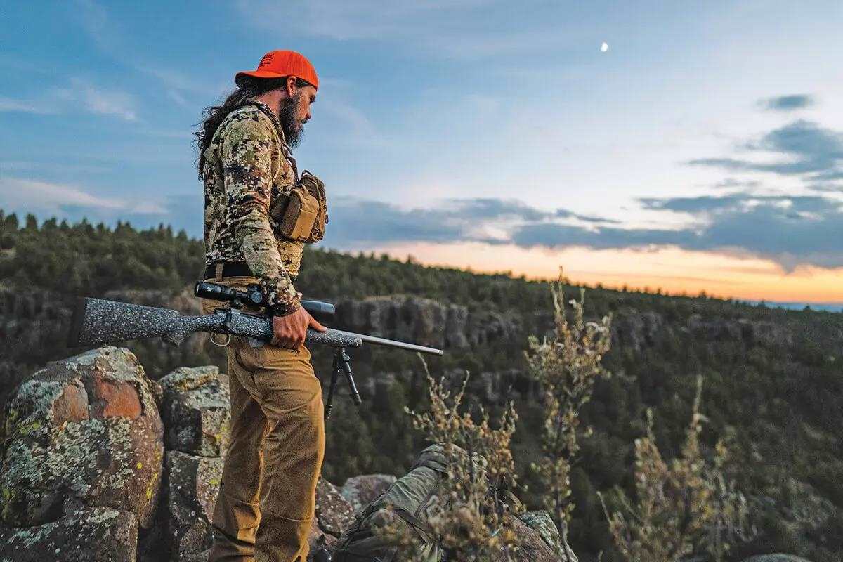 Hunter with long hair, in brown camo and orange ballcap, holding rifle in rocky desert terrain