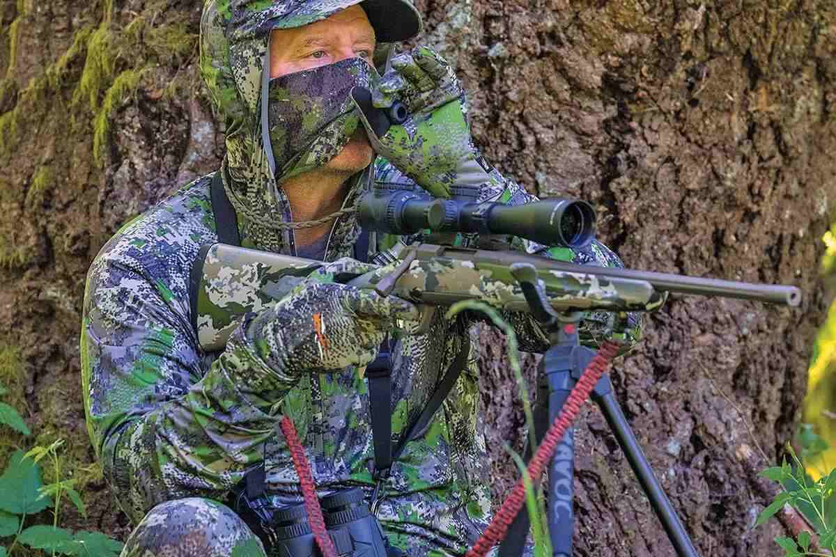 Man in camo head-to-toe in front of tree trunk with riflescope
