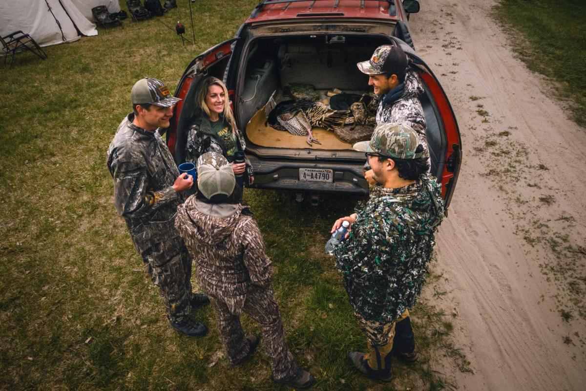 5 hunters in camo standing around back of SUV, where you see recently hunted wild turkeys