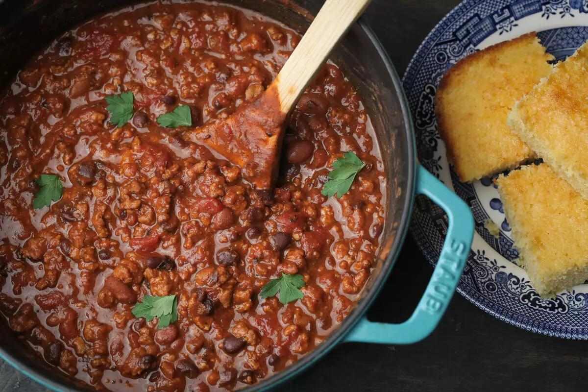 Large cast iron Dutch oven of scrumptious chili with green herbs sprinkled atop and cornbread on a plate next to it