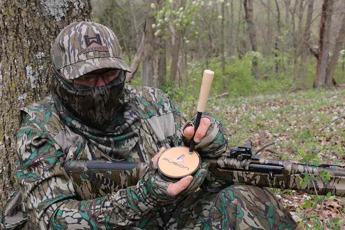 Hunter wearing camo in woods, using turkey call