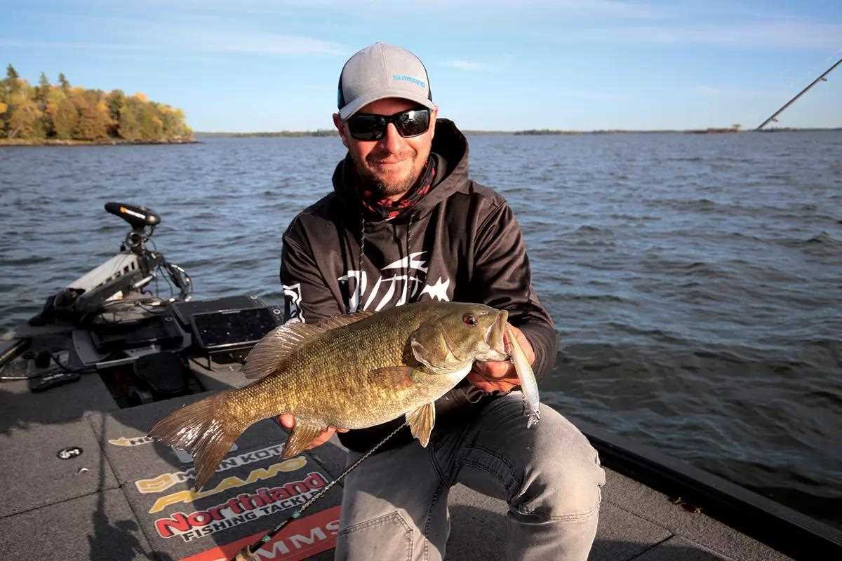 Fisherman in hoodie, ballcap, and shades holding large bass on boat over still waters 