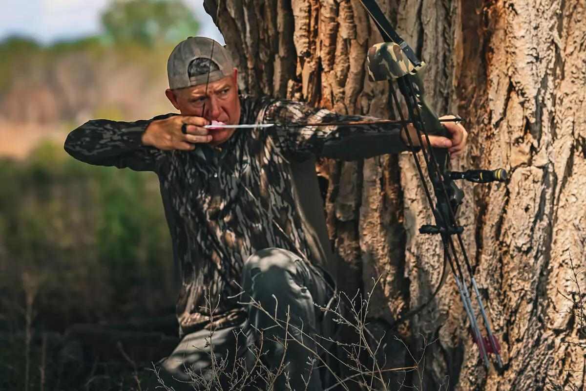 Bowhunter in backwards ballcap and camo taking aim with bow & arrow beside a tree