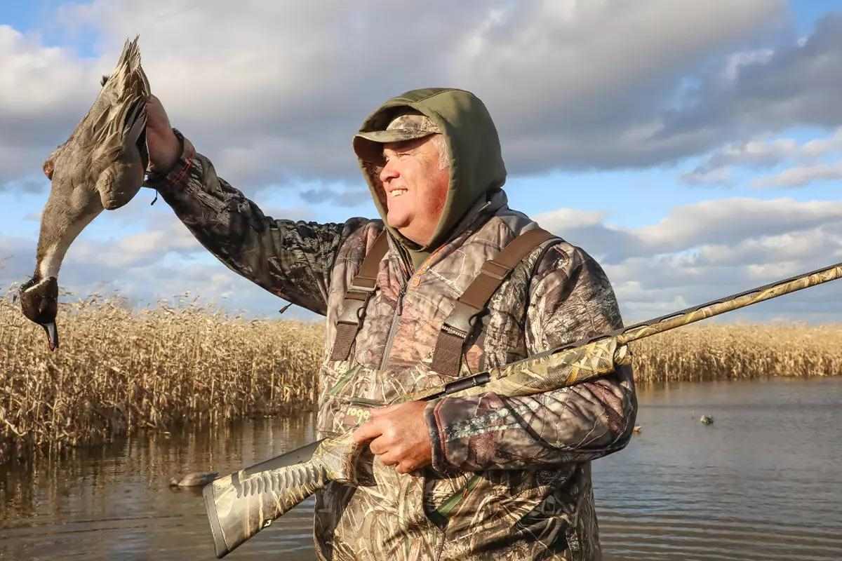 Hunter in camo, holding shotgun and downed waterfowl in front of water