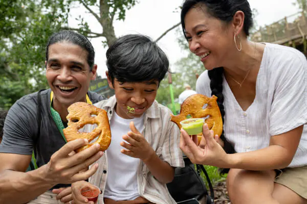 Two parents laughing with their child as they share two Shrekzel pretzels with green cheese at Universal Studios Florida.