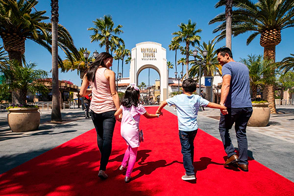 A family standing on a red carpet walks happily towards the arch entrance of Universal Studios Hollywood.