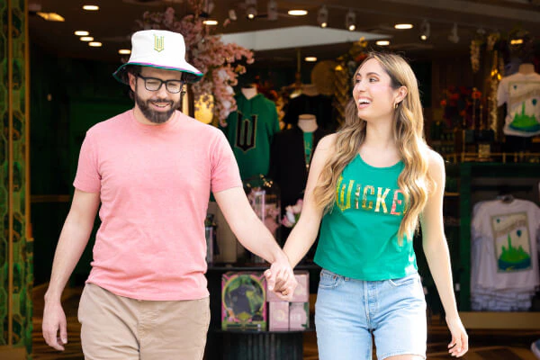 Two guests holding hands while wearing Wicked themed merchandise including a tank top and bucket hat.