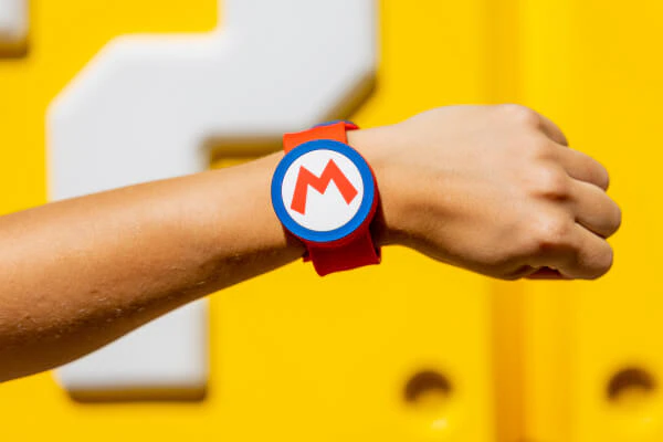 A red, blue and white Power-Up Band™ with “M” for Mario on a guest’s wrist, with yellow question-mark blocks in the background.