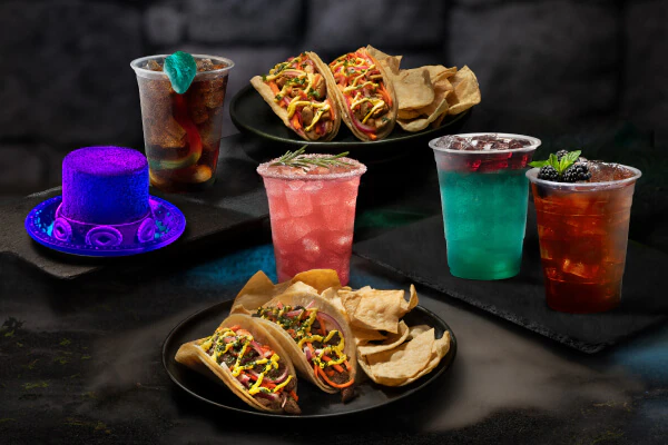 Two black plates of tacos and tortilla chips surrounded by cups of different-colored drinks and a decorative purple top hat.