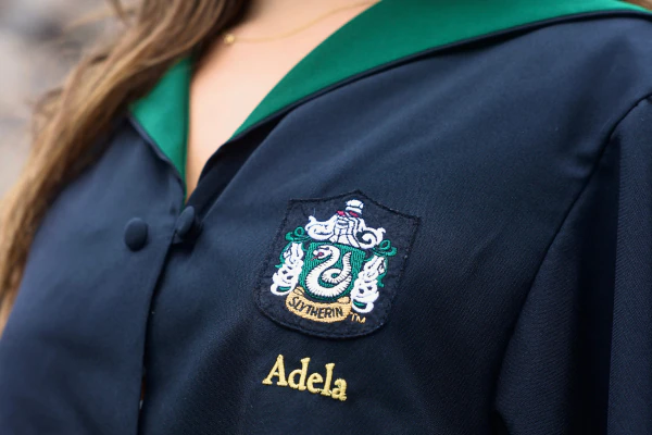 A person wearing a Slytherin robe with an embroidered patch of the house crest and the name Adela underneath.