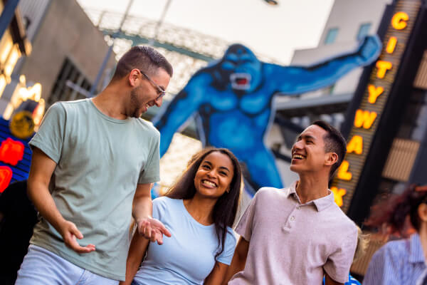 Three friends laughing together with a large blue King Kong figure in the background hanging off a neon “CITYWALK” sign.