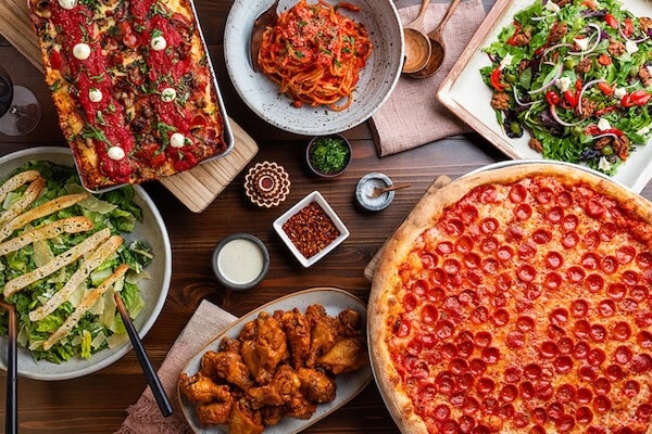 A tabletop featuring two pizzas, chicken wings, spaghetti, two salads and dipping sauces from Slice House.