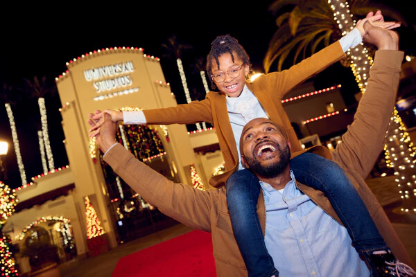 A child sitting on top of their parent's shoulders as they stand in front of the Universal Studios Hollywood arches.