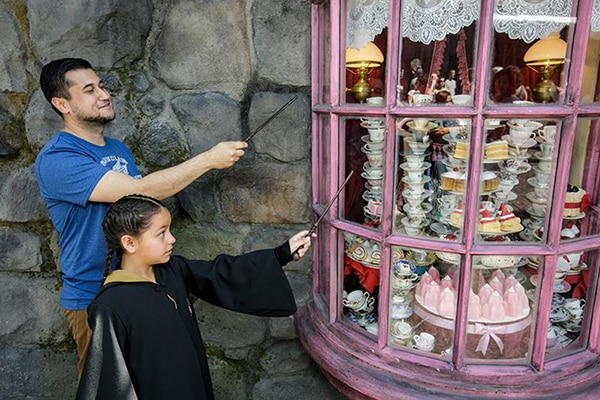 A father standing next to his daughter as they point wands at a storefront window in The Wizarding World of Harry Potter.