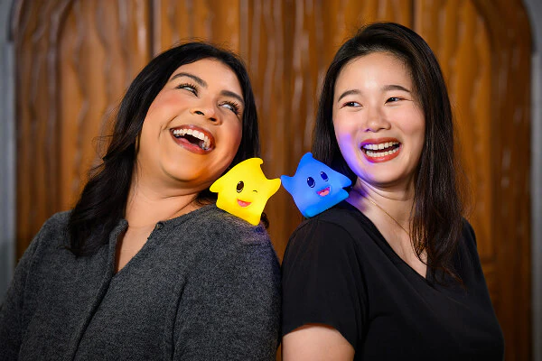 Friends standing back-to-back laughing as the yellow and blue Luma figures on their shoulders interact with each other.
