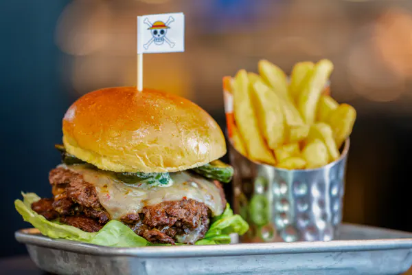 A stacked cheeseburger with green toppings and a ONE PIECE skull-and-crossbones flag next to a side of fries.
