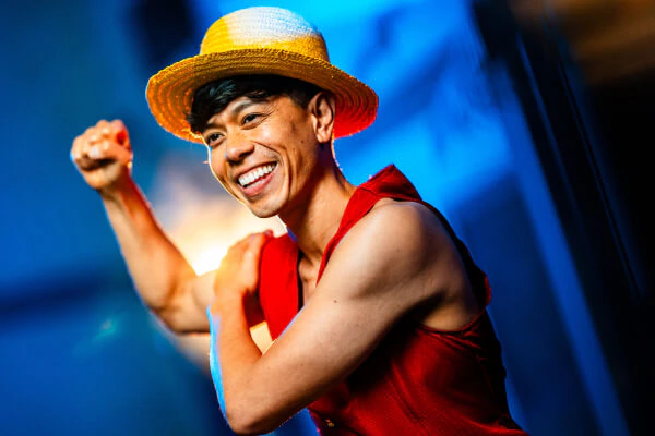 A young man dressed as Luffy from One Piece pumps his fist and smiles.