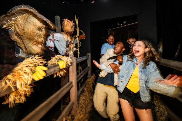 Friends recoiling in fear as they encounter straw scarecrow-like scareactors with burlap-covered heads behind a wooden fence.