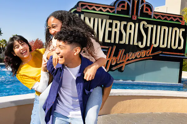 Three friends clinging together and laughing next to the iconic neon Universal Studios Hollywood sign and fountain.