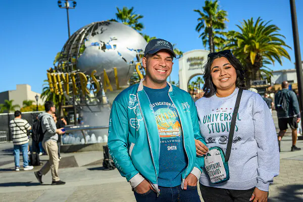 Two guests wearing Universal Studios Hollywood apparel and smiling in front of the Universal Studios globe fountain.