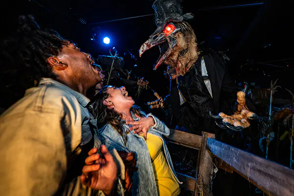 Friends shrieking in horror as a tall, black-suited scareactor with a bird beak, glowing red eyes and a hat confronts them.