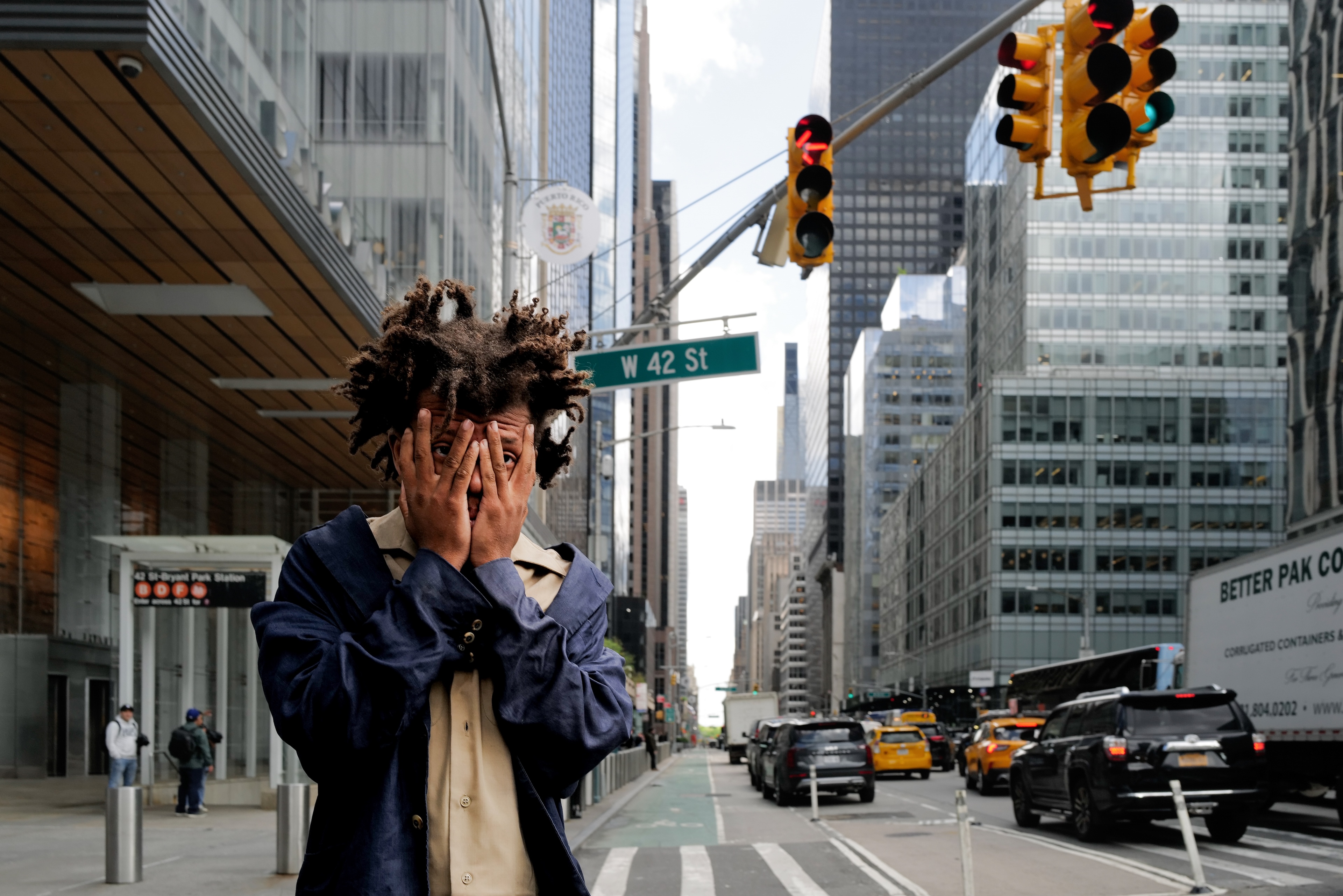 A street scene in NYC. A man peers at us through his fingers. In the background people wait at a bus stop, cars taxis and trucks zoom by.