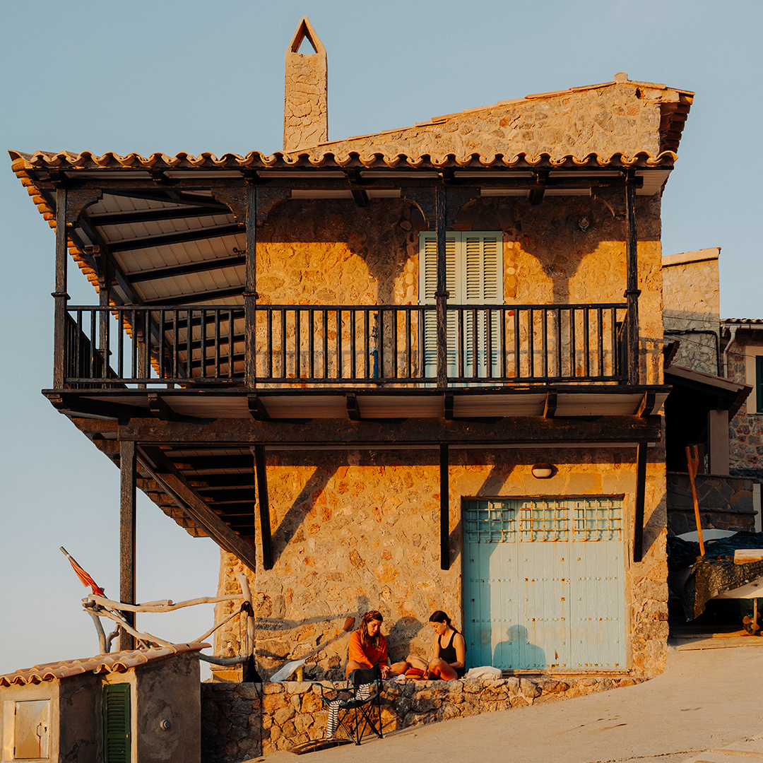 A photograph of a stone house, with slanted roof, pointed chimney, and a wraparound balcony against a clear blue sky. Two people sit in front, bathed in golden hour sunlight.