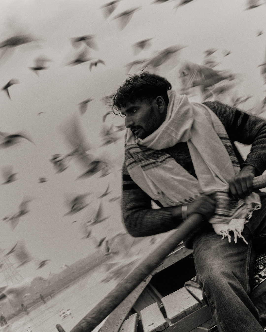 Black and White photograph of a man rowing a boat. Our horizon is tilted to 45 degrees. the man wears a scarf and looks out of frame. Behind him, the blurred chaos of a flock of birds in flight.