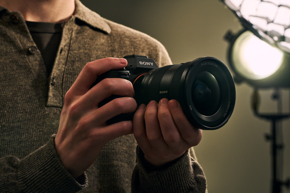 The New Sony A7R Mark VI, held against a persons shoulder. in the background, slightly defocused, two lights and modifiers on stands