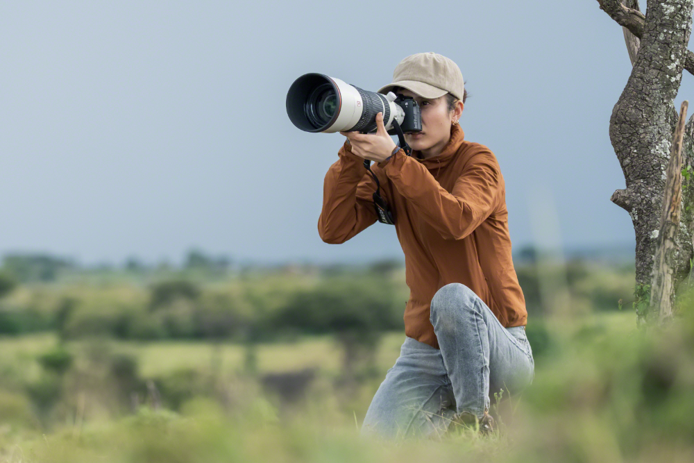 A person in a ball cap kneels in a field, raising a camera to their eye with a large telephoto lens. behind them, a tree. Grasses out of focus in the extreme foreground