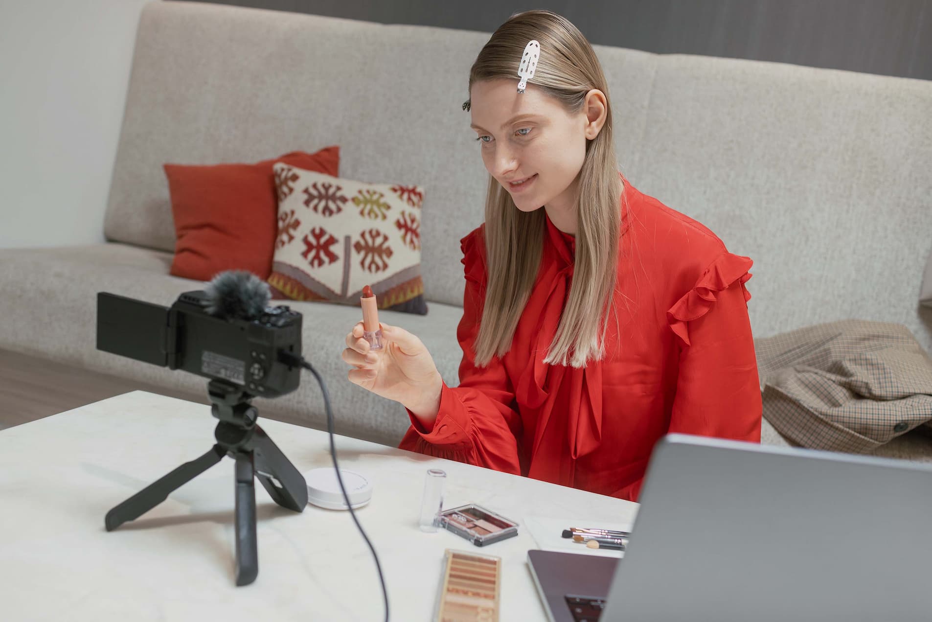 A woman sits on the floor between a couch and coffee table. She is part way through a presentation, holding make up to the camera on a small tripod in front of her